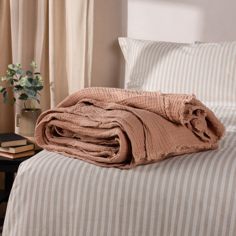 Lark Plain Cotton Throw in pink clay, folded with a crinkled muslin texture and frayed edge, styled on a striped bed beside pillows.