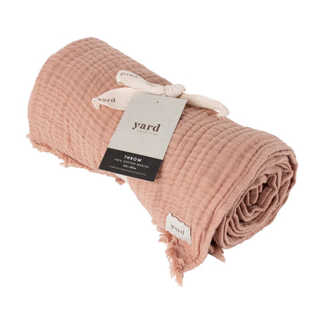 Lark Plain Cotton Throw in pink clay, rolled with crinkled muslin texture and fringed edges, tied with a Yard branded tag, shown on a white background.