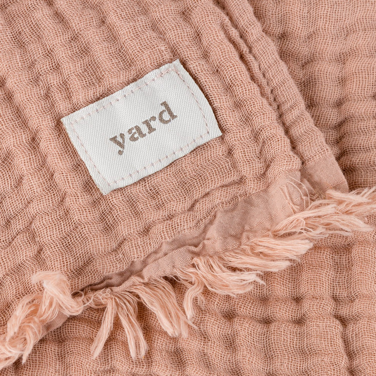 Close-up of the Lark Plain Cotton Throw in pink clay, showing crinkled muslin texture, a woven "yard" label, and a fringed edge.