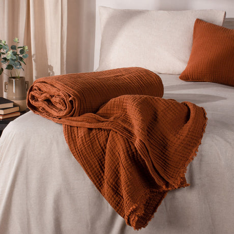 Lark Plain Cotton Throw in pecan, partly rolled and draped across a bed with crinkled cotton muslin texture and a frayed raw edge, styled with cream bedding and a matching cushion.