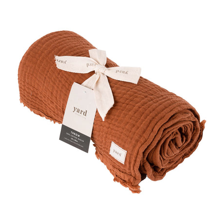 Lark Plain Cotton Throw in pecan, rolled to show its crinkled cotton muslin texture and frayed edge, tied with a cream ribbon and Yard branded tag, on a white background.
