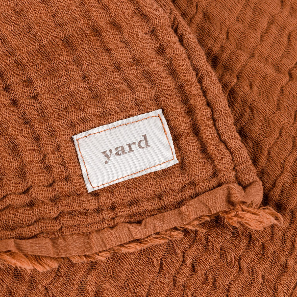 Close-up of the Lark Plain Cotton Throw in pecan, showing crinkled muslin texture, a frayed raw edge, and a woven "yard" fabric label.