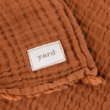 Close-up of the Lark Plain Cotton Throw in pecan, showing crinkled muslin texture, a frayed raw edge, and a woven "yard" fabric label.