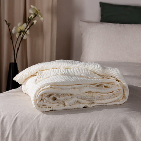 Lark Plain Cotton Throw in white, folded with crinkled muslin texture and frayed edges, shown on a bed beside linen pillows in a neutral bedroom setting.