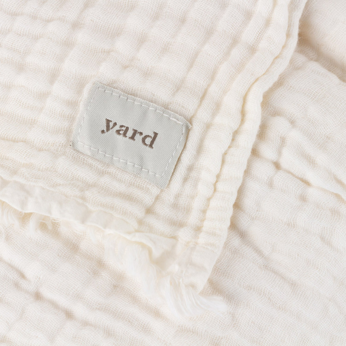 Close-up of the Lark Plain Cotton Throw in white, showing crinkled muslin texture, a frayed edge, and a woven "yard" label stitched to the corner.