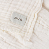 Close-up of the Lark Plain Cotton Throw in white, showing crinkled muslin texture, a frayed edge, and a woven "yard" label stitched to the corner.