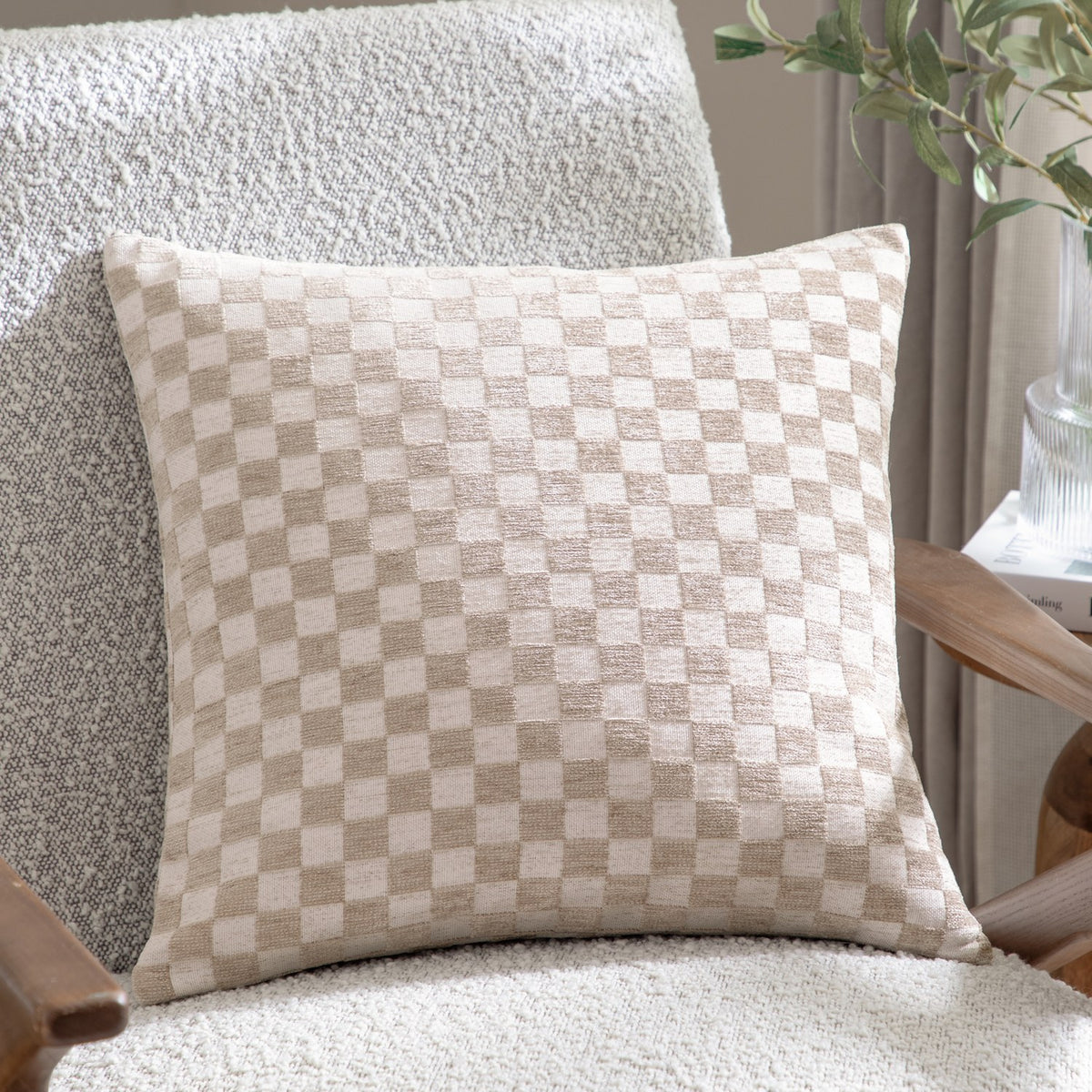 Larro Jacquard Chenille Cushion in Natural, showing a woven checkerboard pattern in beige and cream tones, styled on a bouclé armchair beside a wooden side table.