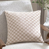 Larro Jacquard Chenille Cushion in Natural, showing a woven checkerboard pattern in beige and cream tones, styled on a bouclé armchair beside a wooden side table.