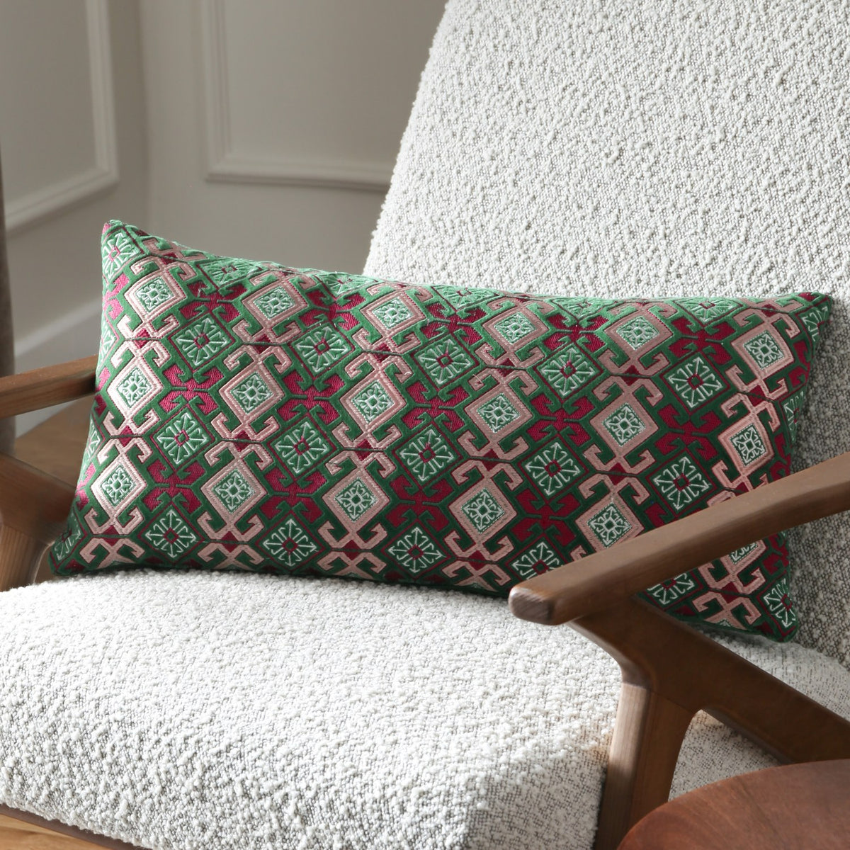 Laurel Velvet Cotton Cushion in Cerise/Jade, a bolster-shaped cushion with embroidered geometric diamond pattern in green, cream and pink, styled on a bouclé armchair.