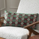 Laurel Velvet Cotton Cushion in Cerise/Jade, a bolster-shaped cushion with embroidered geometric diamond pattern in green, cream and pink, styled on a bouclé armchair.