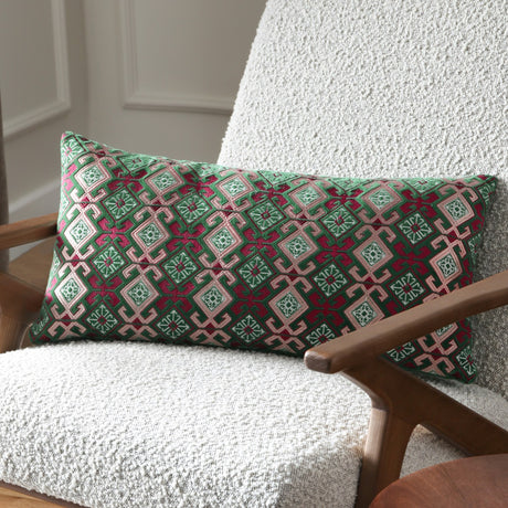 Laurel Velvet Cotton Cushion in Cerise/Jade, a bolster-shaped cushion with embroidered geometric diamond pattern in green, cream and pink, styled on a bouclé armchair.