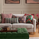 Laurel Velvet Cotton Cushion in Cerise/Jade, a bolster-shaped cushion with embroidered geometric diamond pattern, shown on a beige sofa styled with patterned cushions in a living room setting.