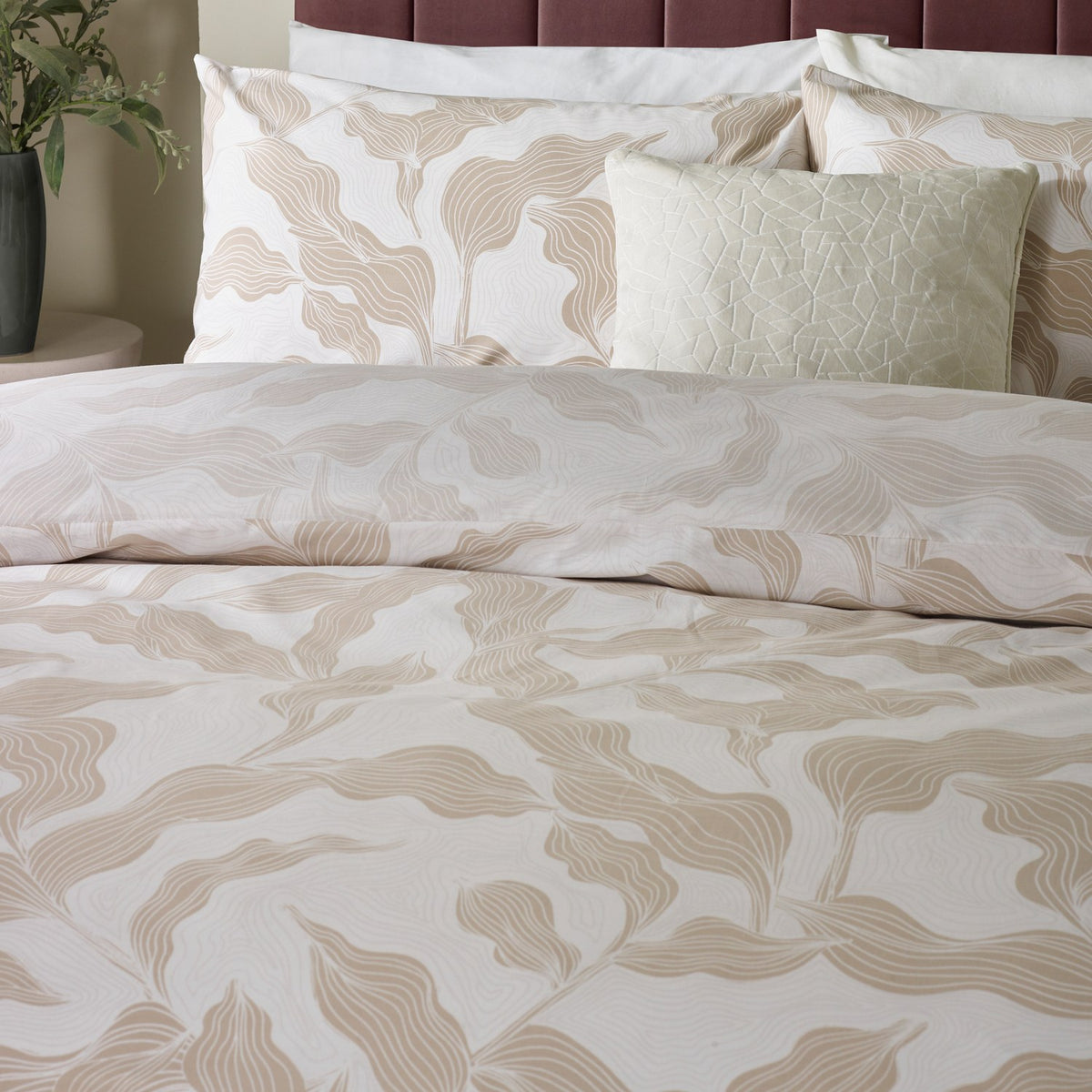 Lauro Reversible Leaf Duvet Cover Set in Stone, styled on a bed with matching pillowcases and cushions, showing a linear leaf print in soft tan and cream tones.