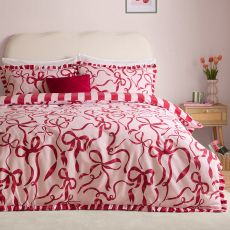 La Vie En Bows duvet cover set on a bed with pink and red bow-print duvet and pillowcases, edged in red and white striped ruffle trim, in a soft pink bedroom.