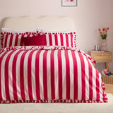 La Vie En Bows reversible duvet cover set in red and pink candy stripe on a bed with ruffled edges and matching pillowcases, styled in a bedroom with a cream headboard.