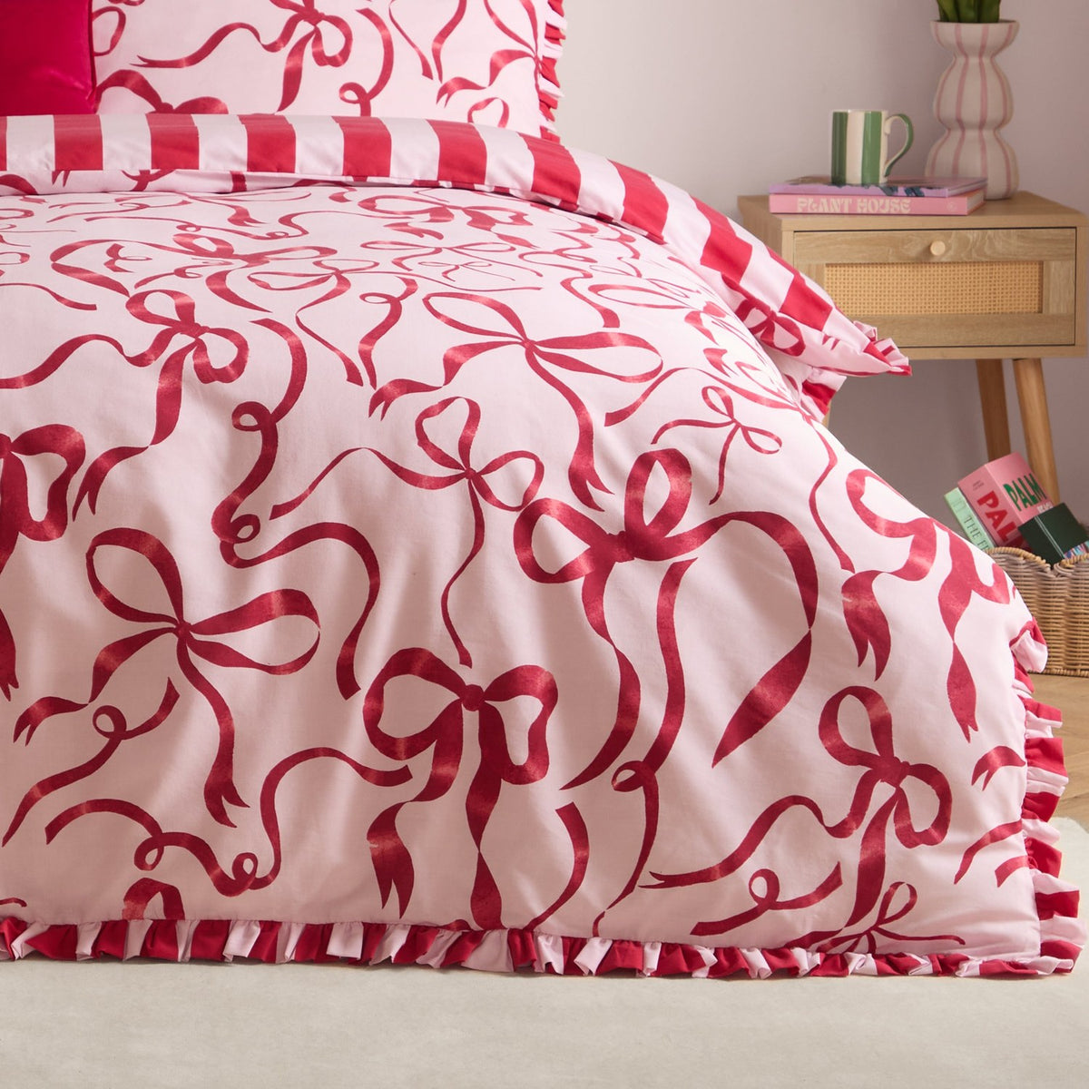 La Vie En Bows duvet cover set on a bed, showing red bow print on pink fabric with a red and white striped ruffle trim and matching pillowcase, in a bedroom setting.