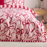 La Vie En Bows duvet cover set on a bed, showing red bow print on pink fabric with a red and white striped ruffle trim and matching pillowcase, in a bedroom setting.
