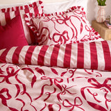 La Vie En Bows reversible duvet cover set on a bed, showing pink fabric with red bow prints alongside matching red and pink candy stripe pillowcases and ruffled trim.