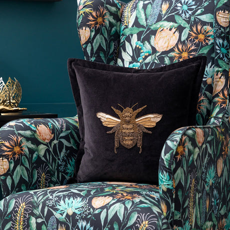 Layla Animal Velvet Cushion in black with gold embroidered bee motif and flat Oxford-style border, shown on a floral upholstered armchair.