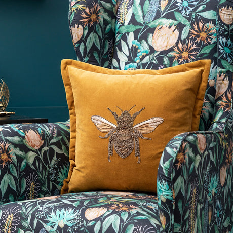 Layla Animal Velvet Cushion in mustard, a square velvet cushion embroidered with a gold beaded bee design, shown on a floral-print armchair.