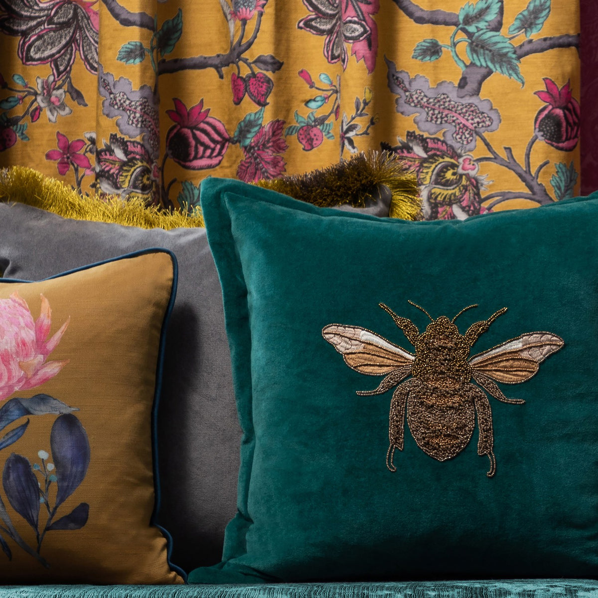 Layla Animal Velvet Cushion in teal with a beaded bee motif in amber and gold thread, shown among patterned cushions and a floral curtain backdrop.