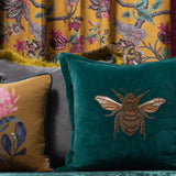 Layla Animal Velvet Cushion in teal with a beaded bee motif in amber and gold thread, shown among patterned cushions and a floral curtain backdrop.