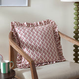 Lulah Block Floral Linen Cushion in red and natural with ruffled edge trim, styled on a rattan-armed wooden chair beside a green table lamp.