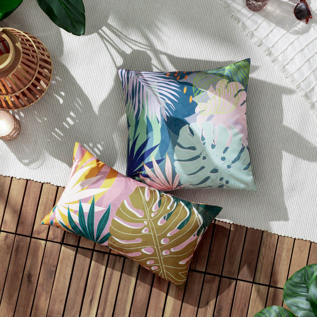 Leafy Reversible Polyester Cushion in teal with pale palm fronds, mint monstera leaves and pink accents, shown with a matching tropical-print lumbar cushion on an outdoor rug.