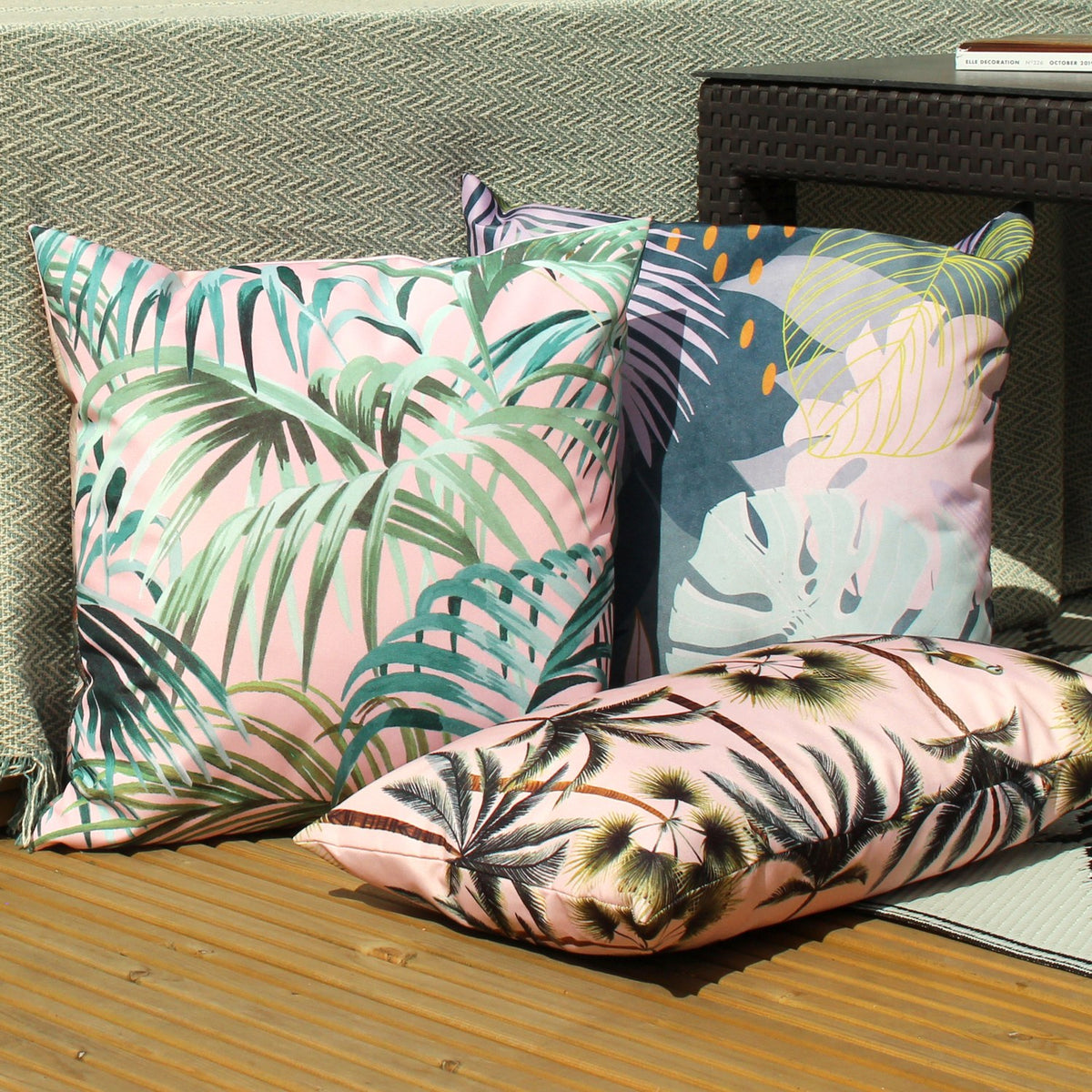 Leafy Reversible Polyester Cushion in Teal shown among pink palm-print cushions, featuring an abstract tropical leaf design with mint monstera and amber dot accents, styled outdoors on a wooden deck.