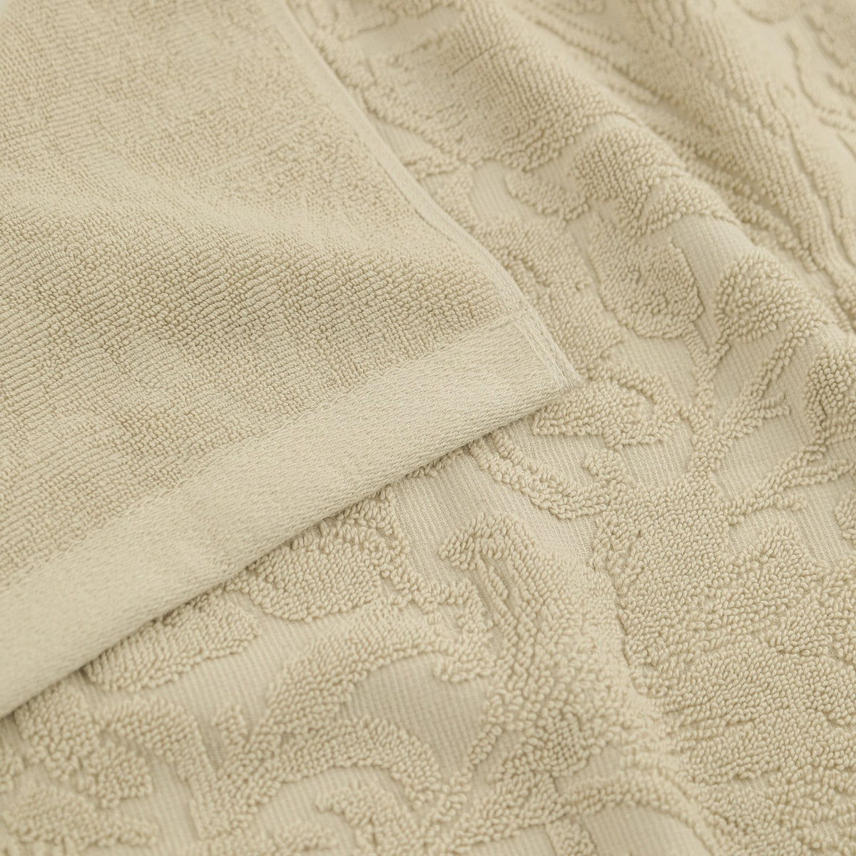Close-up of a Leicester floral cotton towel in natural, showing folded plain-woven edge and a raised scrolling leaf and bloom pattern sculpted into the pile.