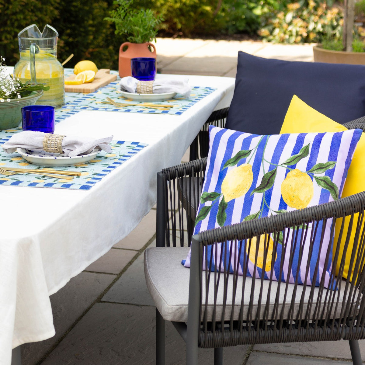 Lemons Abstract Polyester Cushion in blue and lilac stripes with a leafy lemon print, styled on an outdoor chair beside a garden dining table.