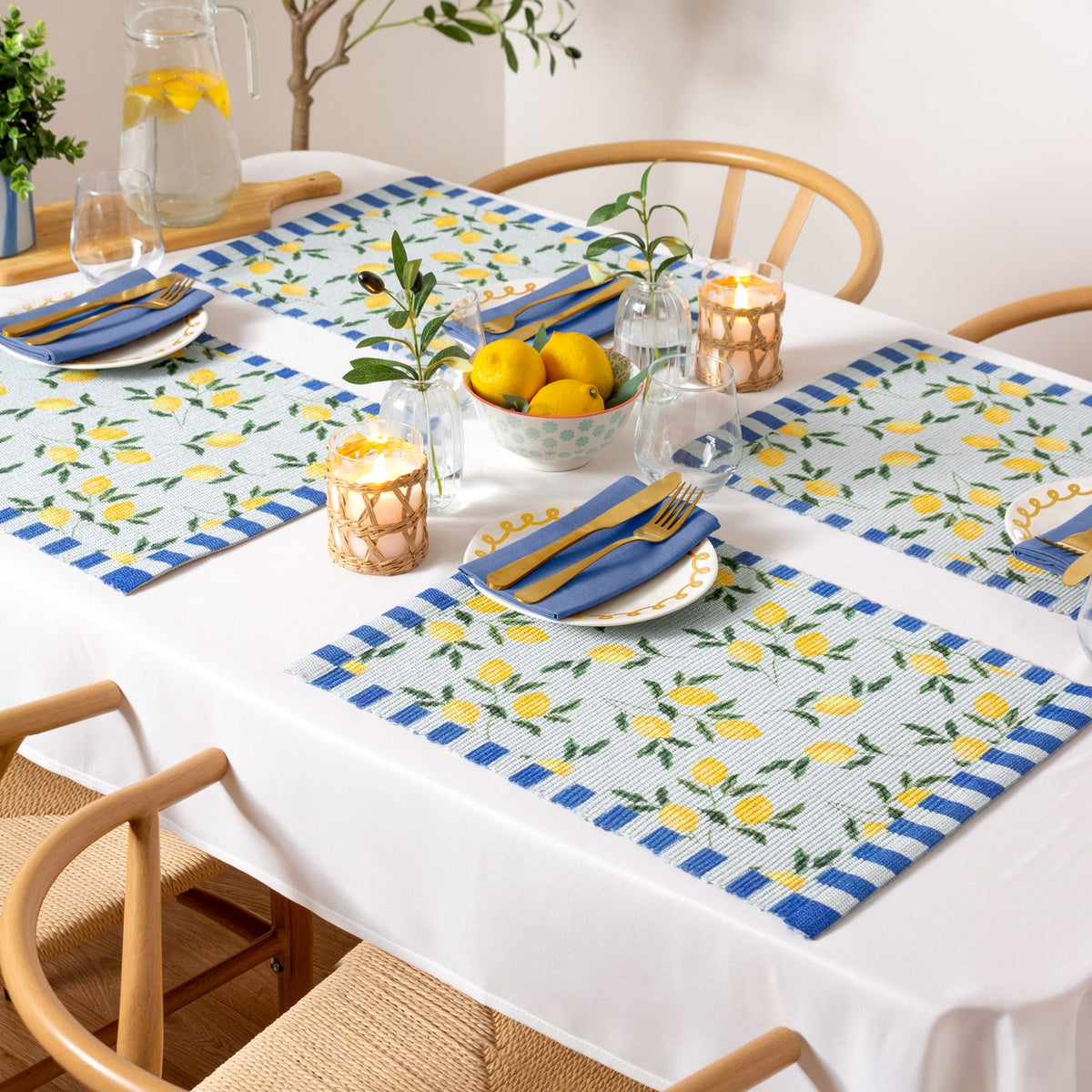 Lemons Floral Polyester Placemats in blue set on a dining table with striped borders, yellow lemon and leaf print, styled with blue napkins, gold cutlery, candles, and a lemon centrepiece.
