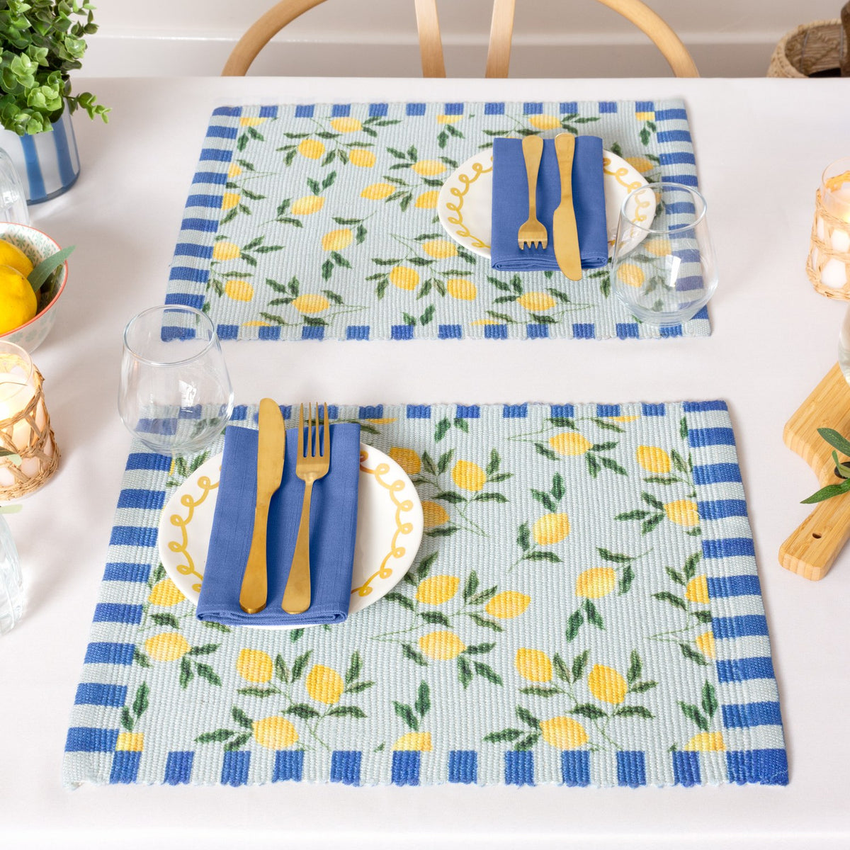 Two Lemons Floral polyester placemats in blue, printed with yellow lemons and green leaves and striped blue borders, set on a dining table with plates, cutlery, and blue napkins.