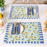 Two Lemons Floral polyester placemats in blue, printed with yellow lemons and green leaves and striped blue borders, set on a dining table with plates, cutlery, and blue napkins.