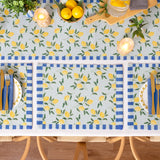 Rectangular polyester placemats with a lemon and leaf print on a pale ground, bordered by blue striped edges, shown styled on a dining table with matching tablecloth and place settings.