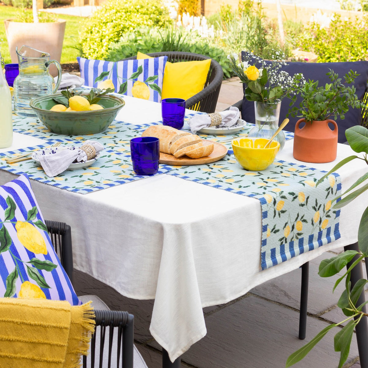 Lemons Floral Polyester Placemats in blue used as a table runner on an outdoor dining table, featuring yellow lemon and green leaf prints bordered by blue stripes.