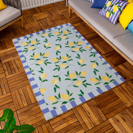 Lemons Striped Polyester Rug in blue, featuring a yellow lemon and leaf pattern on a pale blue ground with a blue and cream block border, shown on a wooden deck beside outdoor sofa cushions.
