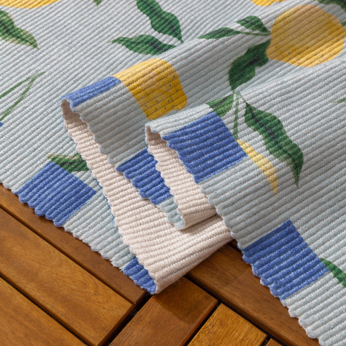 Folded corner of the Lemons Striped Polyester Rug in blue, showing the ribbed weave, lemon and leaf print, and blue-cream block border, resting on a wooden surface.