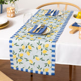 Lemons Floral Polyester Table Runner in blue, laid on a dining table with a lemon and foliage print and blue striped edges, styled with plates, cutlery, and glassware.