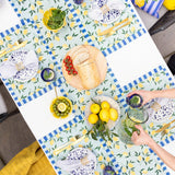Blue and yellow lemon-print polyester table runner styled on an outdoor table with bread, olives, tomatoes, lemons, and a jug of infused water.