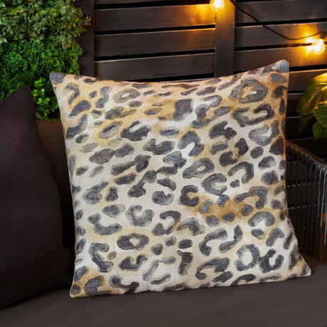 Square Leopardis cushion in woven polyester with a painterly leopard print in charcoal, cream, amber and tan, shown outdoors on a dark sofa.