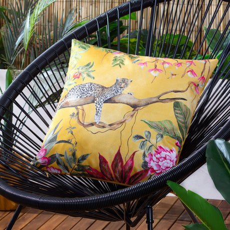 Square gold polyester cushion with a hand-painted leopard on a branch, tropical foliage and pink florals, shown on a black woven outdoor chair.