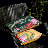 Leopard Jungle Polyester Cushion in gold and green shown on a rattan chair, printed with a leopard on a branch, tropical leaves and pink peonies, outdoor setting.