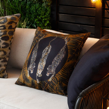 Leopardis reversible polyester cushion, 50x50cm, black and gold, showing three leopards in a dark medallion framed by amber palm fronds, styled on a sofa with a leopard-print cushion.