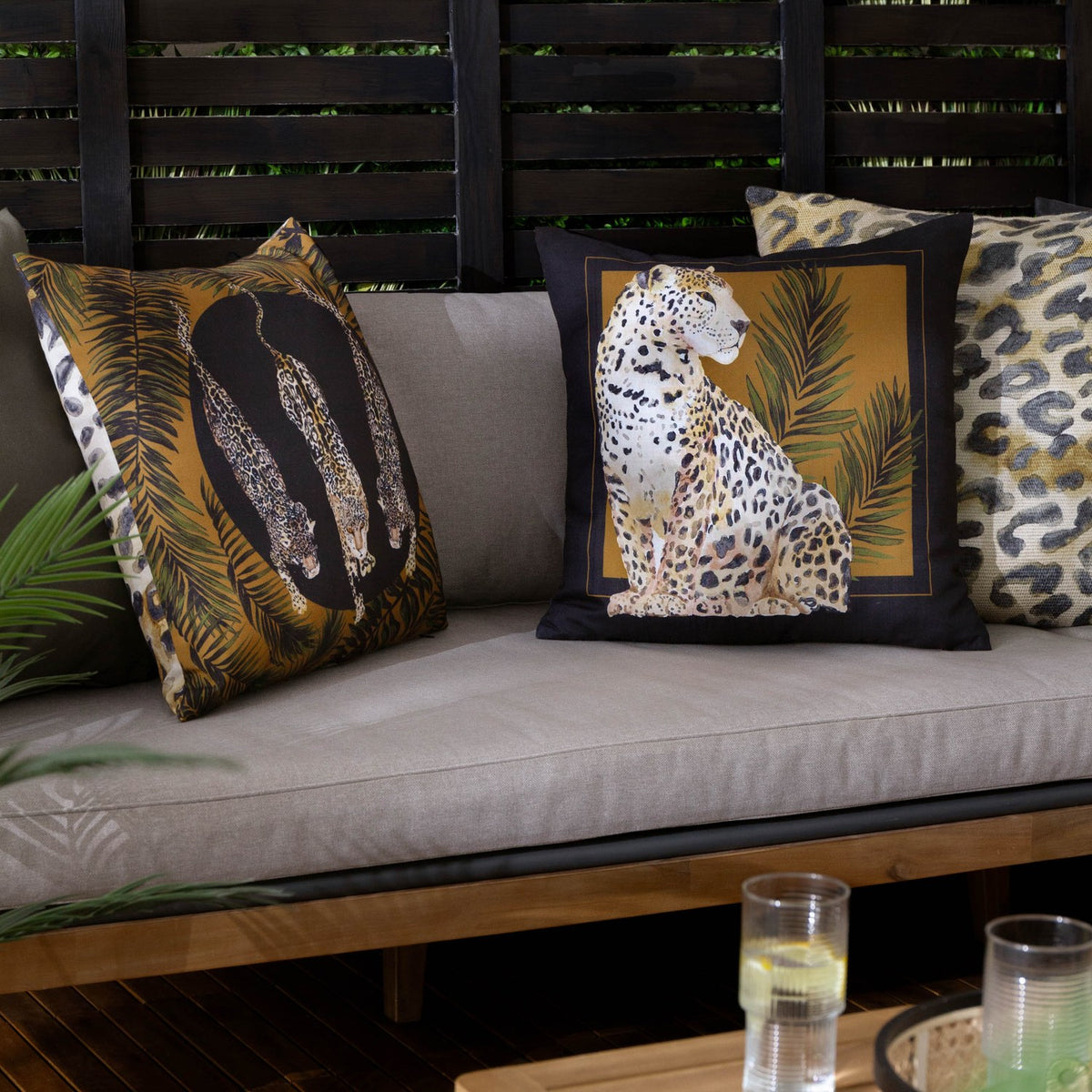 Leopardis reversible polyester cushion in black and gold on an outdoor sofa, showing a printed leopard trio design beside a seated leopard cushion and leopard-print scatter cushions.