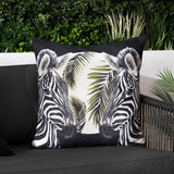 Leopardis reversible polyester cushion in black, cream and olive, featuring two facing zebra heads amid palm fronds, shown on a dark sofa in an outdoor living space.