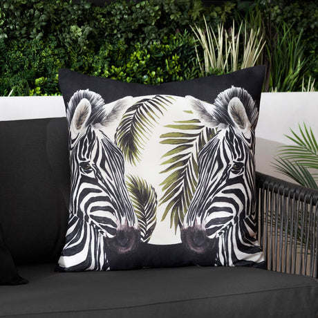Leopardis reversible polyester cushion in black, cream and olive, featuring two facing zebra heads amid palm fronds, shown on a dark sofa in an outdoor living space.
