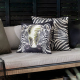 Leopardis reversible polyester cushion in black and cream with a printed zebra-head and palm frond design, styled on a beige outdoor sofa with a zebra-stripe cushion behind it.