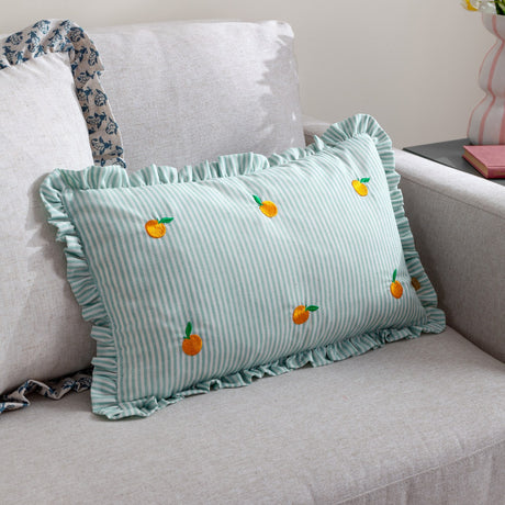 Les Fruites rectangular cushion in green and white pinstripe polyester with ruffle trim and embroidered orange fruit motifs, shown on a light grey sofa.