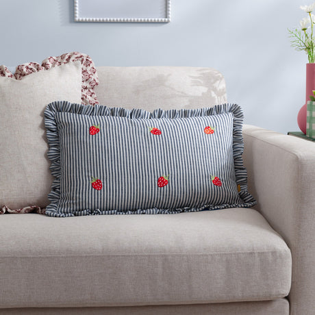 Rectangular blue-and-white pinstriped cushion with embroidered red strawberry motifs and a ruffled edge, styled on a beige sofa in a living room.